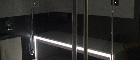 Una sauna moderna, con poca luz, que incluye un panel de vidrio, un banco de madera y un cabezal de ducha metálico.