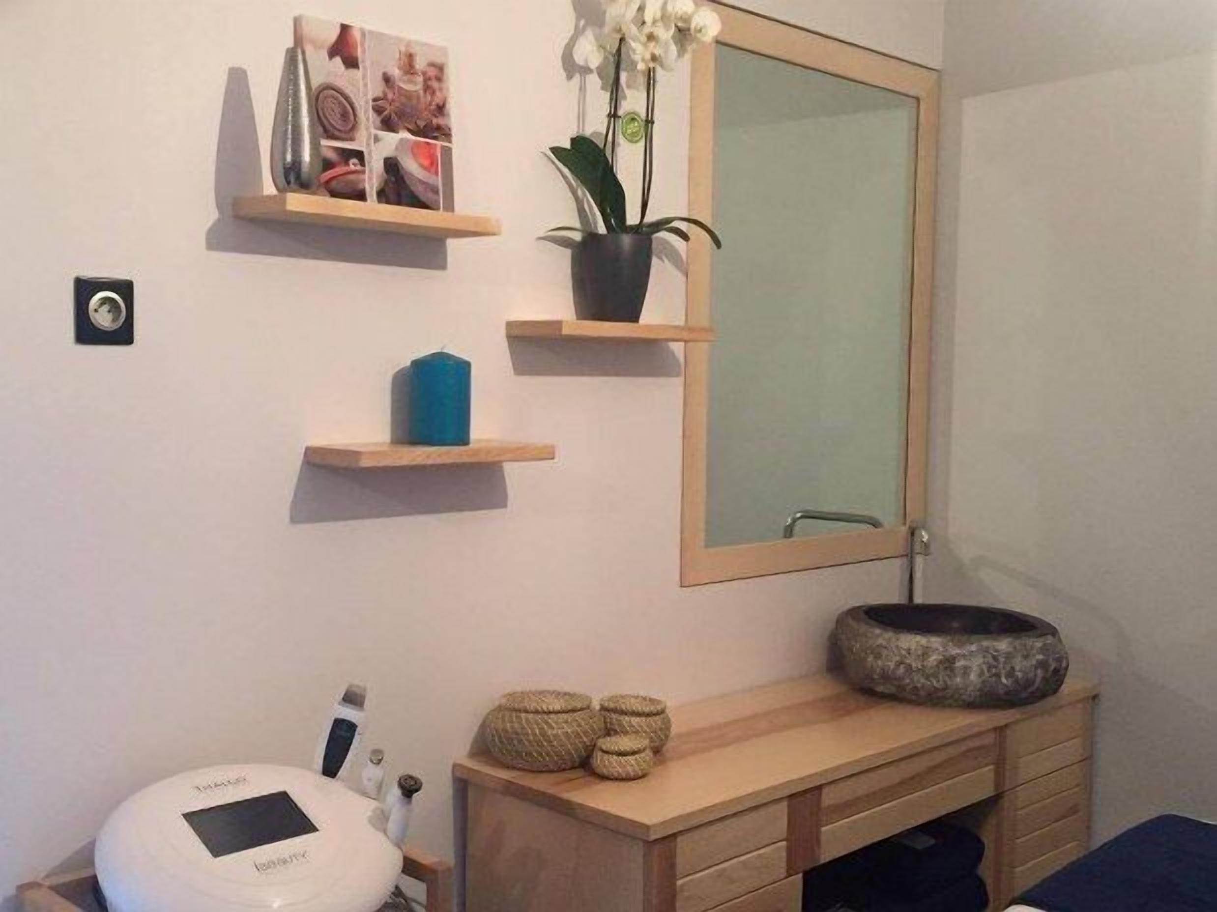 A bathroom with a wooden vanity, a mirror, a small shelf with a plant, a blue candle, and a white device on the counter.
