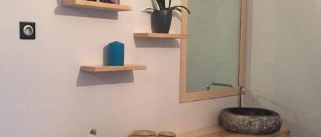 A bathroom with a wooden vanity, a mirror, a small shelf with a plant, a blue candle, and a white device on the counter.