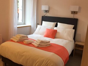 A neatly made bed with white and orange bedding, two bedside lamps, and a window with curtains.