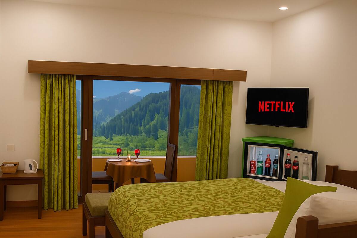 A hotel room with a bed, a table with two glasses and a candle, a TV on the wall, and a view of mountains through the window.
