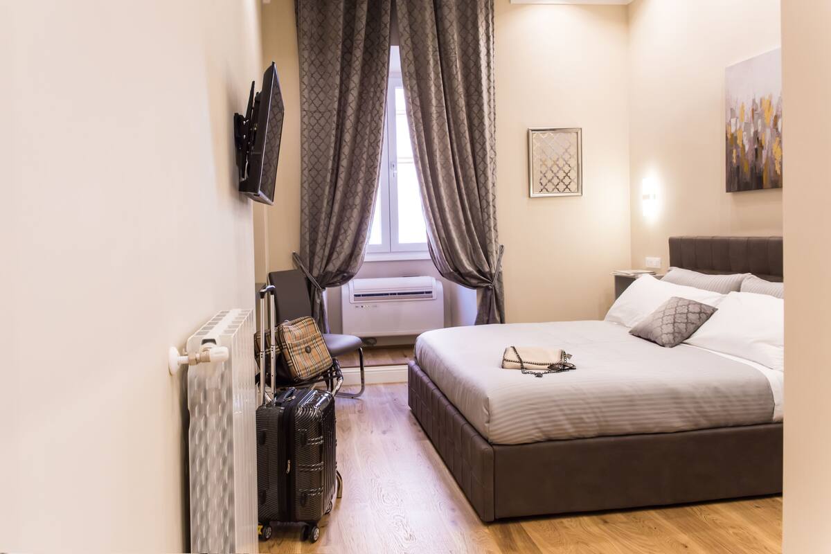 A hotel room with a bed, a suitcase, a chair, and an air conditioning unit.