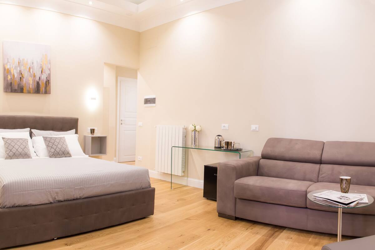 A modern bedroom with a large bed, a sofa, a glass coffee table, and a painting on the wall.