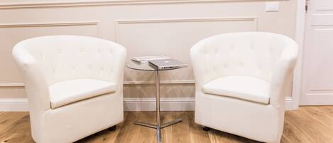 Twee witte fauteuils met een klein tafeltje ertussen, een houten vloer en een witte deur op de achtergrond.
