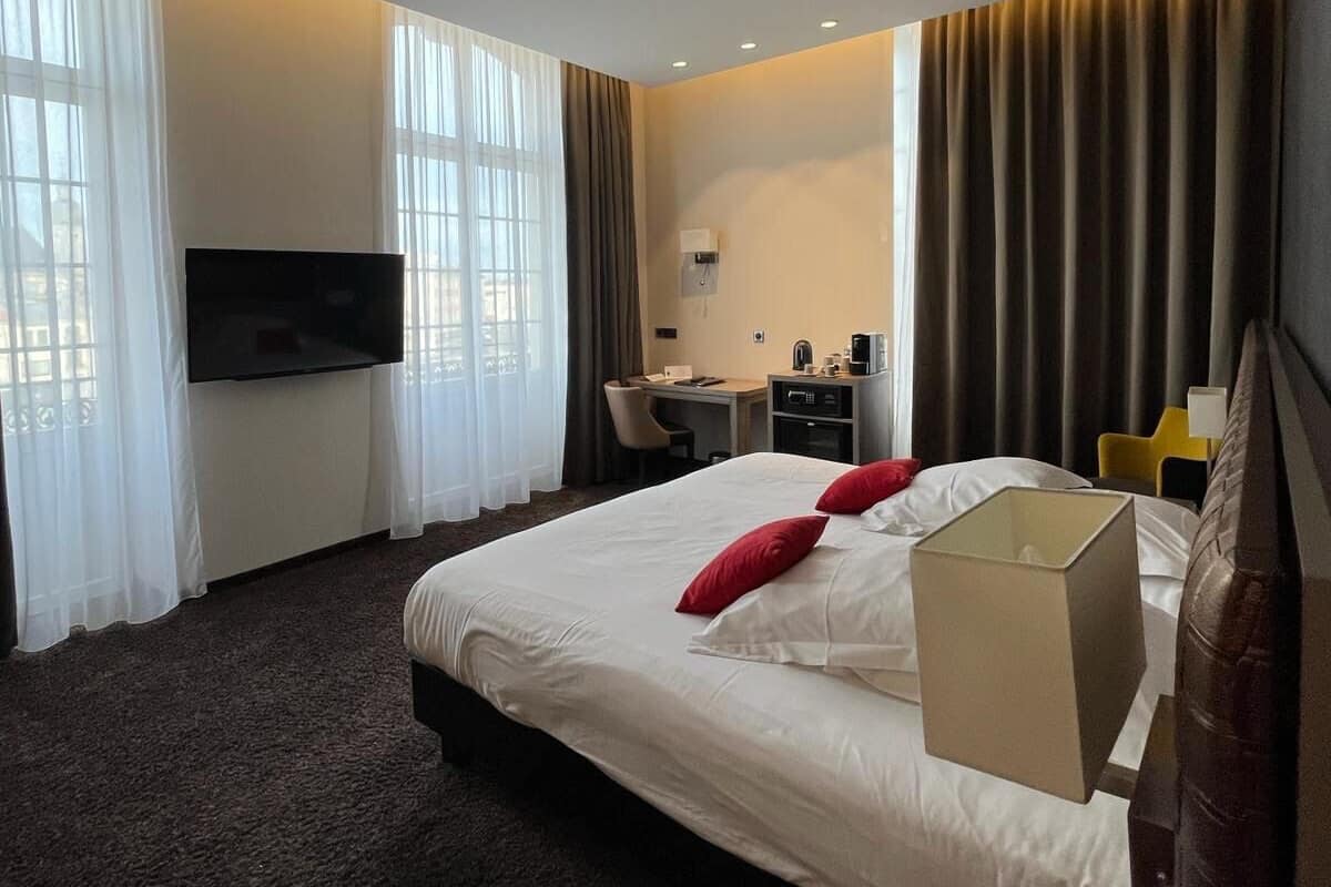Ein Hotelzimmer mit einem großen Bett, einem Schreibtisch mit Stuhl, einem an der Wand befestigten Flachbildfernseher und einem Fenster mit Vorhängen.