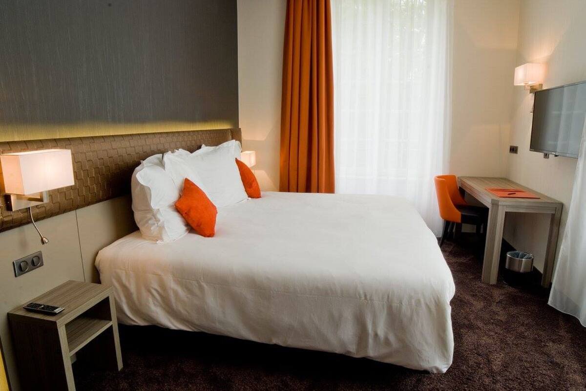 Ein Hotelzimmer mit einem großen Bett, zwei Nachttischlampen, einem Schreibtisch mit Stuhl und einem an der Wand befestigten Fernseher.