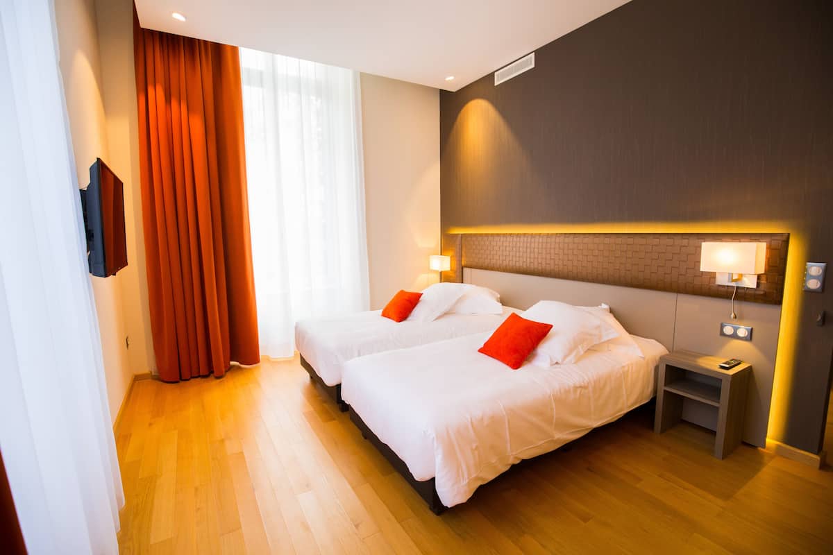 Ein Hotelzimmer mit zwei Betten, einem Nachttisch, einer Lampe und einem Fenster mit Vorhängen.