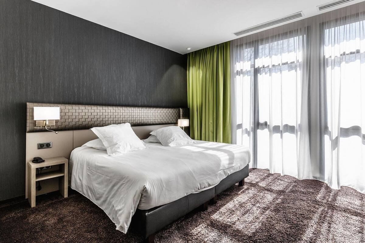 Ein modernes Hotelzimmer mit einem großen Bett, einem Nachttisch, einer Lampe und Blick ins Freie durch transparente Vorhänge.