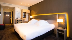 A hotel room with a large bed, bedside tables, a wardrobe, and a seating area with a red ottoman.