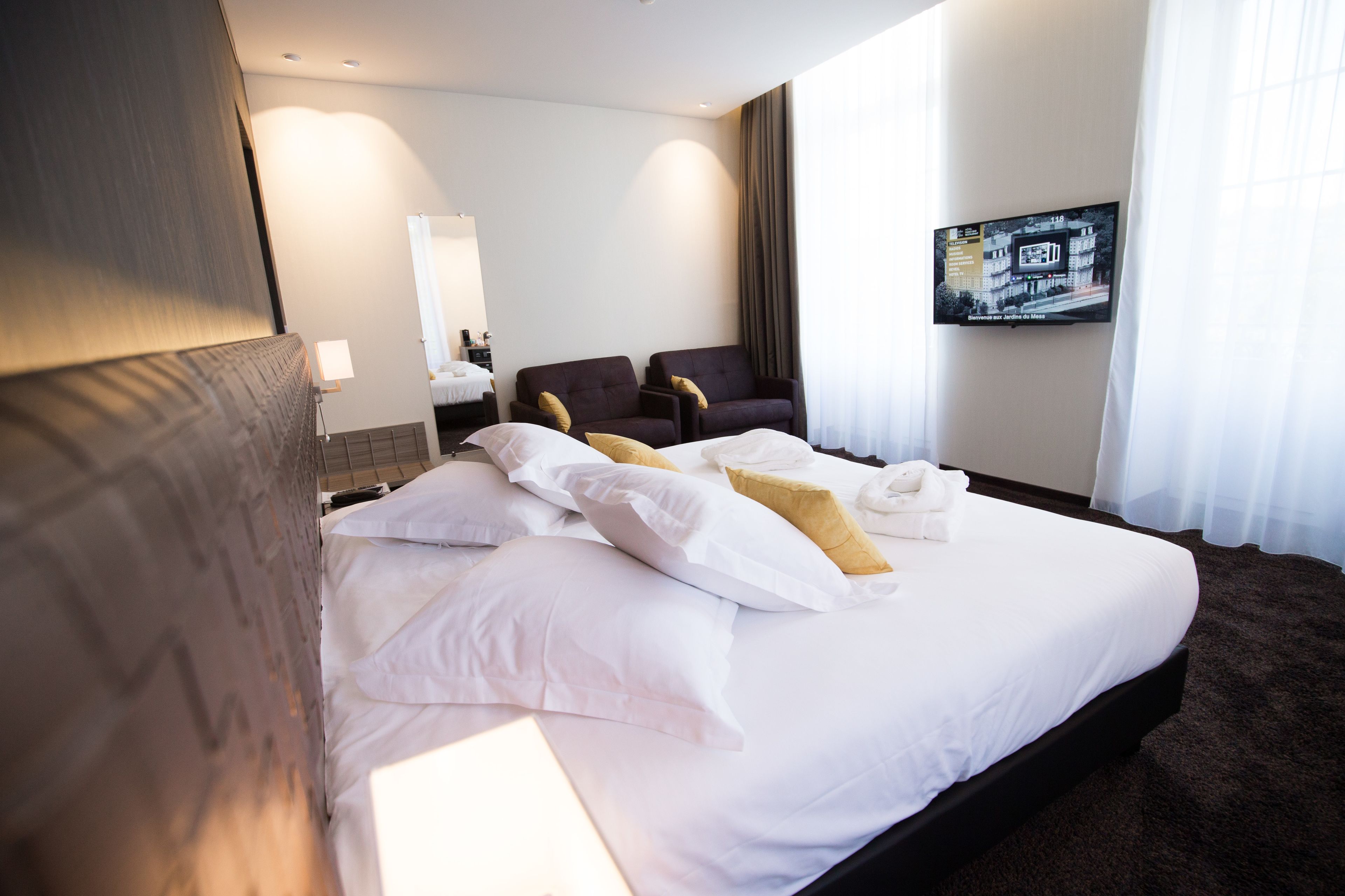A hotel room with a large bed, two armchairs, a TV on the wall, and a window with curtains.