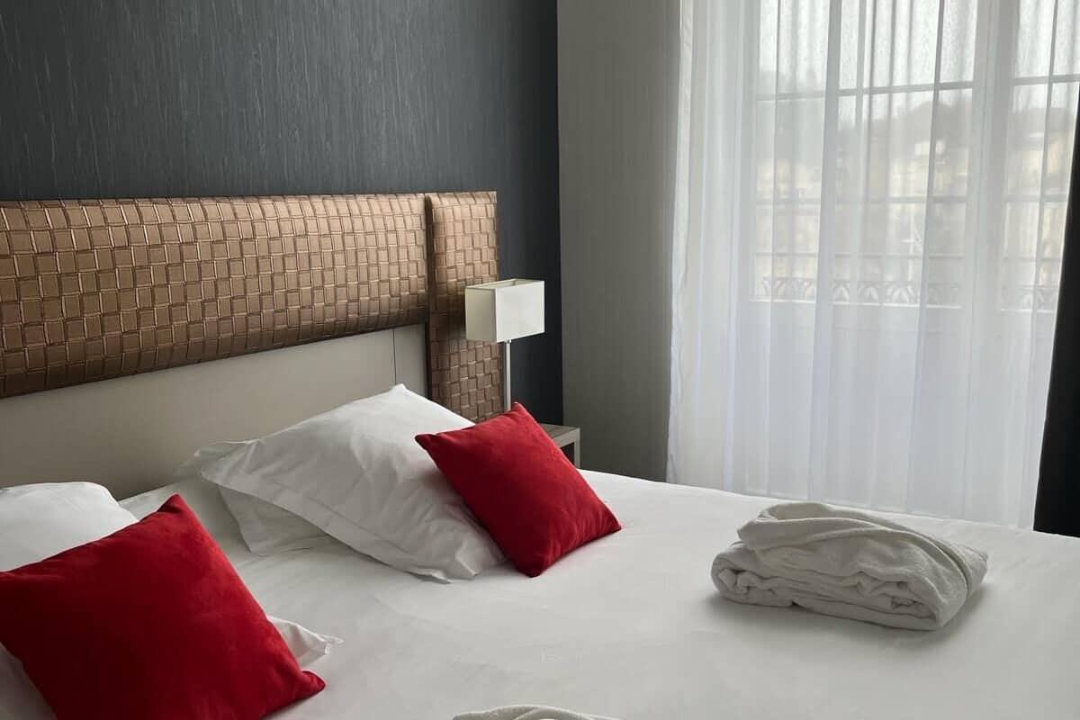 Ein Hotelzimmer mit einem großen Bett, einem Ziegelstein-Muster am Kopfteil, weißer Bettwäsche, roten und weißen Kissen, einer weißen Lampe und einem Fenster mit transparenten Vorhängen.