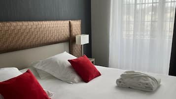 Habitación de hotel con una cama grande, cabecera con diseño de ladrillo, ropa de cama blanca, almohadas rojas y blancas, una lámpara blanca y una ventana con cortinas de gasa.