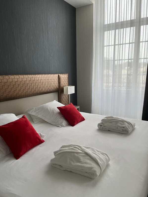A hotel room with a large bed, a headboard with a brick pattern, white bedding, red and white pillows, a white lamp, and a window with sheer curtains. - Les Jardins du Mess (Verdun)