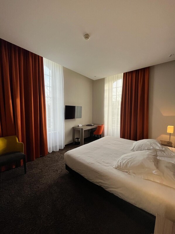 A hotel room with a large bed, a desk with a chair, a television, and curtains. - Les Jardins du Mess (Verdun)
