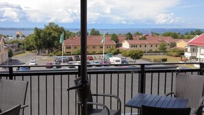 Un balcon avec des fauteuils en osier et une table, donnant sur une rue bordée de voitures garées et de maisons.