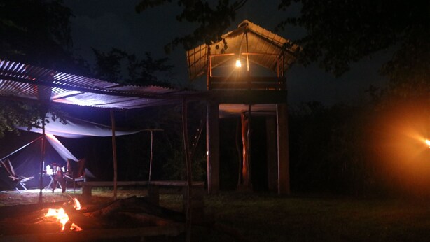 A gazebo with a roof, a small wooden structure, and a campfire.