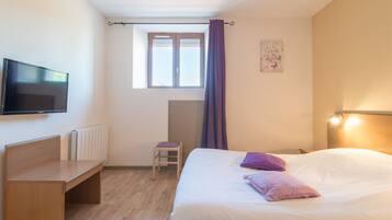 Quarto de hotel com cama, escrivaninha, cadeira, televisão, janela com cortinas e radiador.