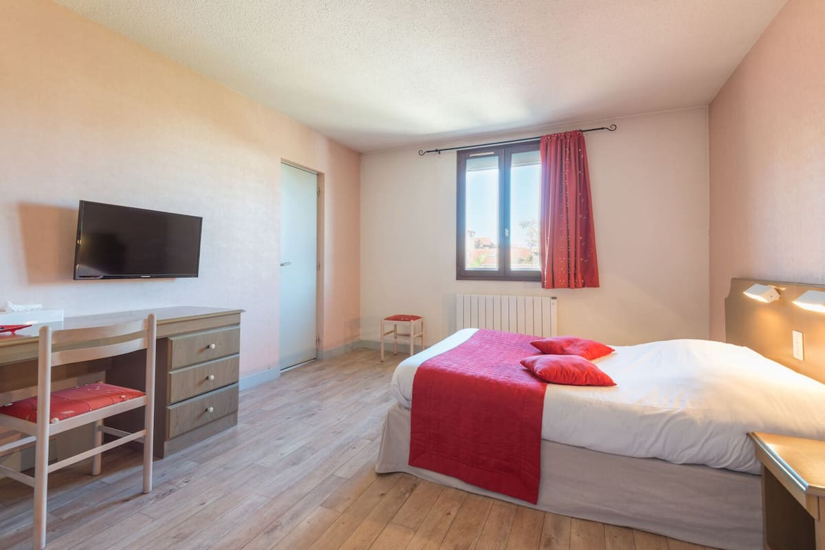 A bedroom with a bed, a desk, a chair, a TV, and a window with curtains.