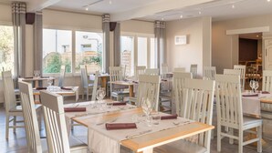 A dining area with multiple tables and chairs, large windows with curtains, and a view of the outside. - Le Réverbère - La Cuisinerie - L'Hôtellerie (Vic-en-Bigorre)