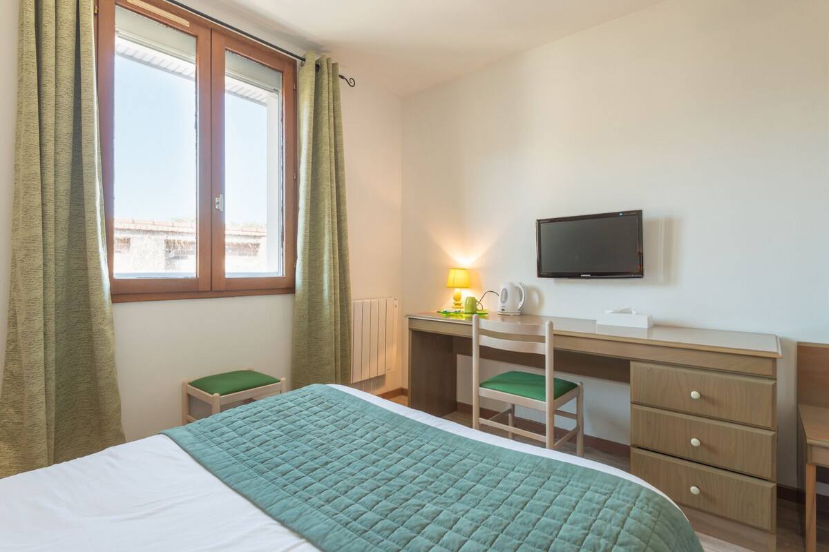 A hotel room with a bed, a desk with a chair, a television, and a window with curtains.