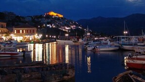 Une ville côtière de nuit, avec des bateaux amarrés dans le port, une colline illuminée et une mer calme qui reflète les lumières.
