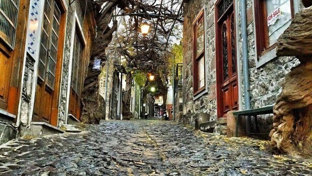 Une ruelle étroite pavée, bordée de bâtiments en pierre, dont certains sont dotés de volets en bois et d’enseignes, qui mène à un point de vue lointain.