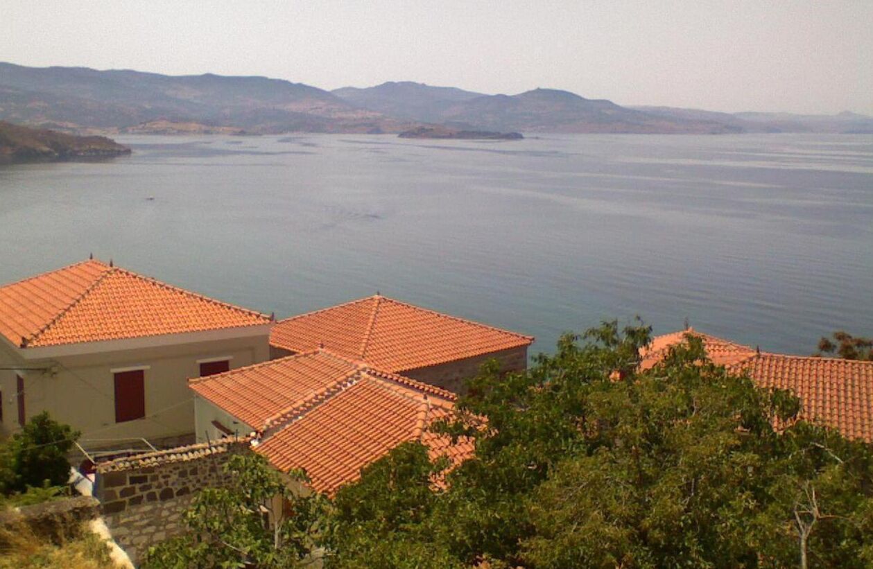 Vista costera con edificios de tejados de terracota que dan sobre un mar tranquilo y colinas lejanas.