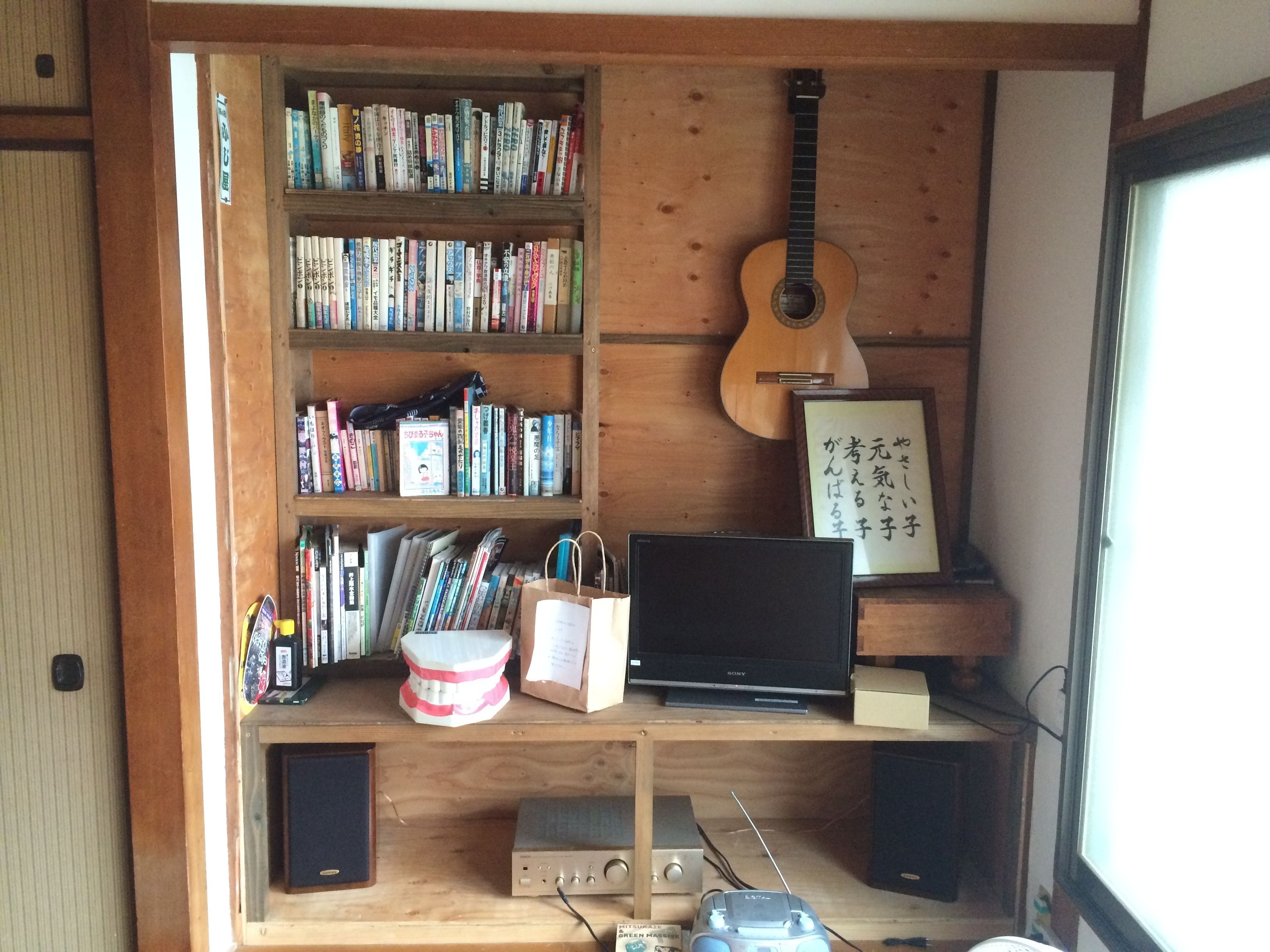 Una habitación con estanterías de madera llenas de libros, una guitarra, un cuadro con texto japonés, un televisor Sony y equipo de sonido.