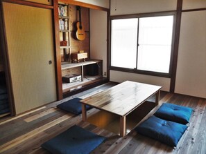 A room with a wooden floor, a table, blue cushions, a bookshelf, a guitar, and a sliding door. - FUJIYA GUEST HOUSE - Hostel (Matsuyama)