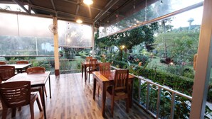 Un espace de restauration en plein air avec des tables et des chaises en bois, une rambarde en verre et une vue sur un jardin luxuriant.