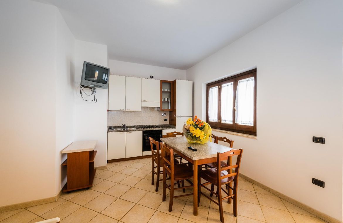 A compact kitchen with white cabinets, a dining table with chairs, a TV mounted on the wall, and a window providing natural light.