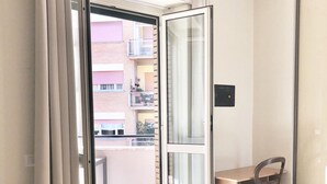 Ein Hotelzimmer mit Bett, Stuhl, Tisch und Blick auf einen Balkon sowie umliegende Gebäude.
