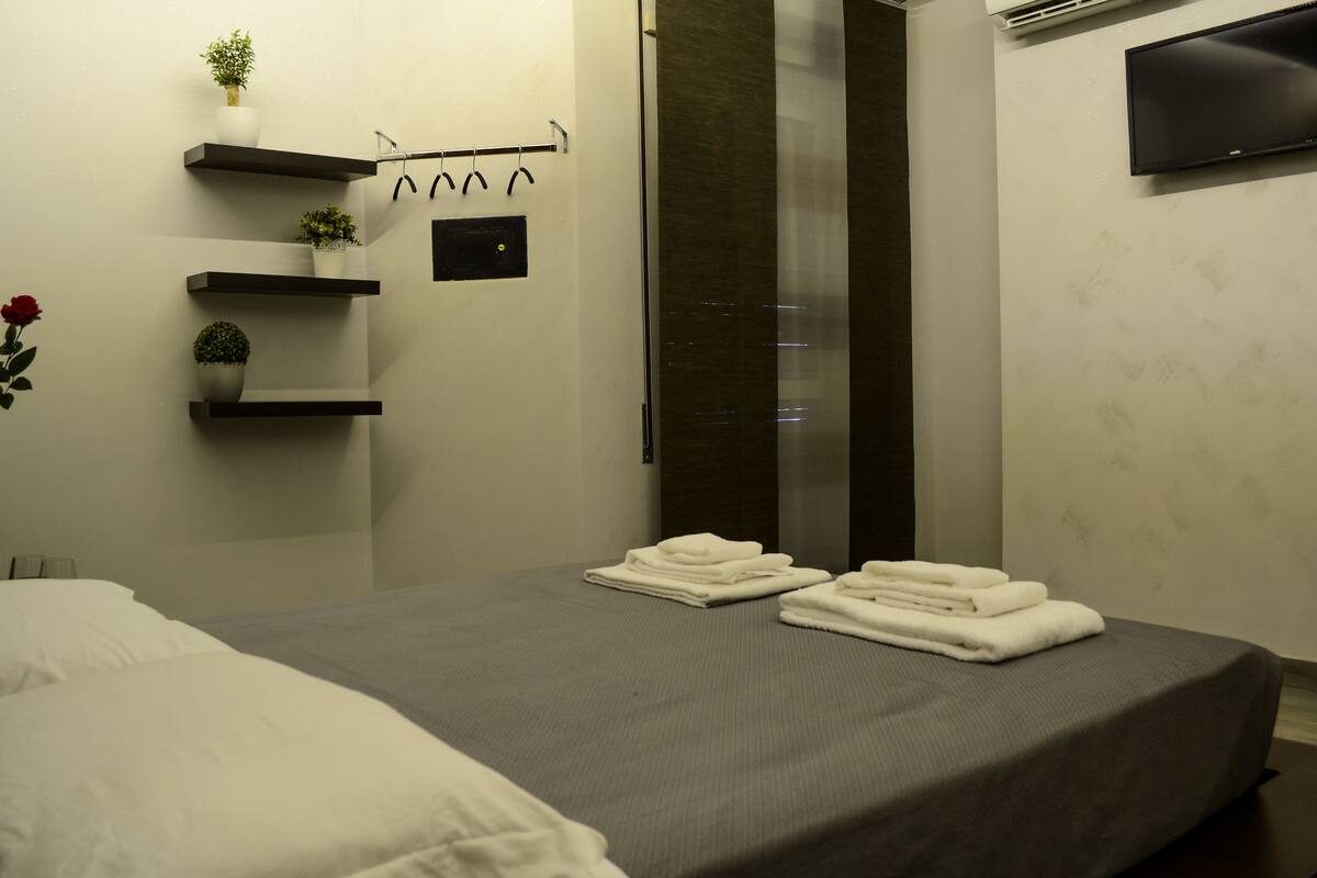 A neatly made bed with folded towels, a potted plant on a shelf, and a flat-screen TV mounted on the wall.
