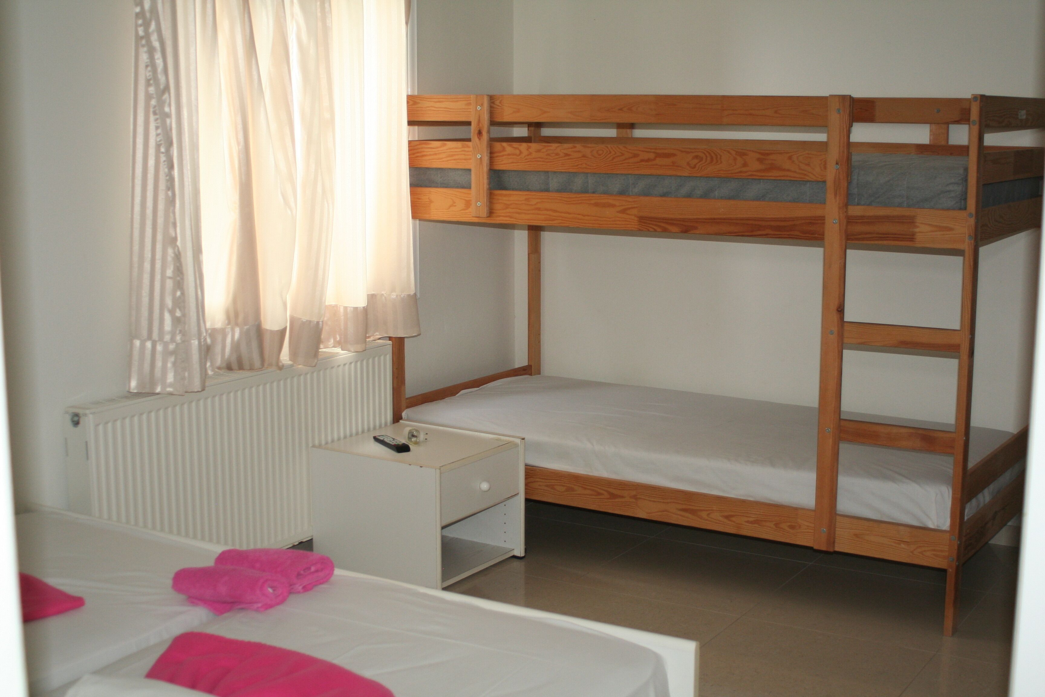 A bunk bed with a white nightstand and a small drawer, a white radiator, and a window with sheer curtains.