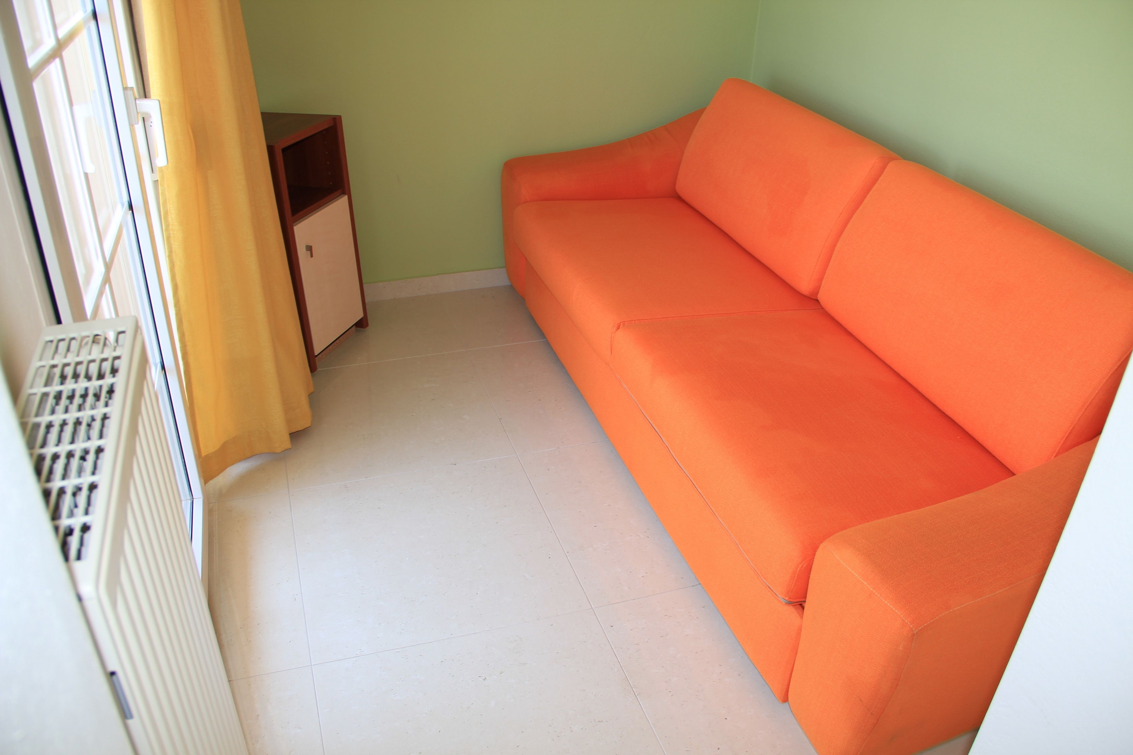 A room with a white radiator, a small wooden cabinet, and an orange sofa against a green wall.