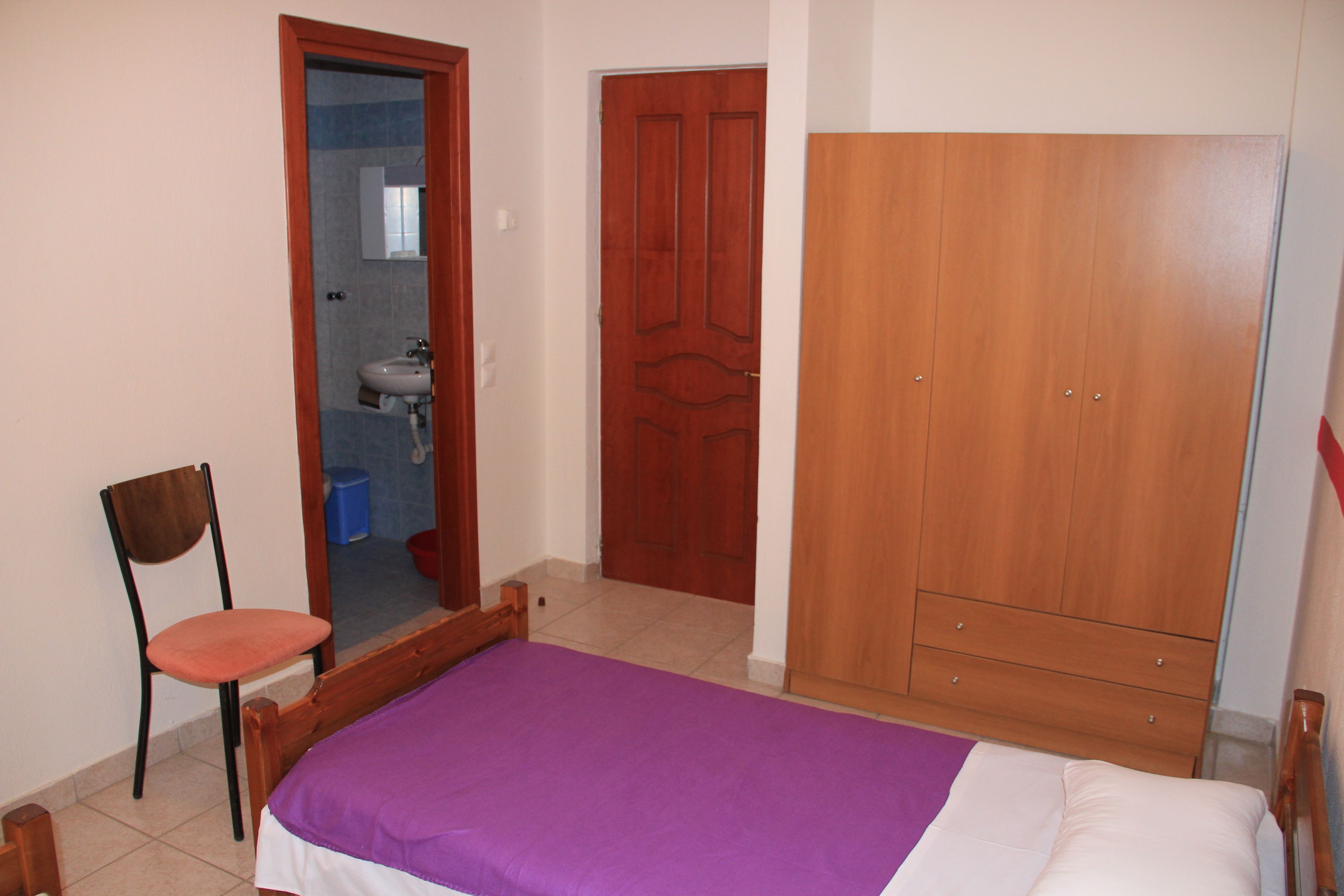 A bedroom with a bed, a wooden wardrobe, a chair, and a bathroom visible through an open door.