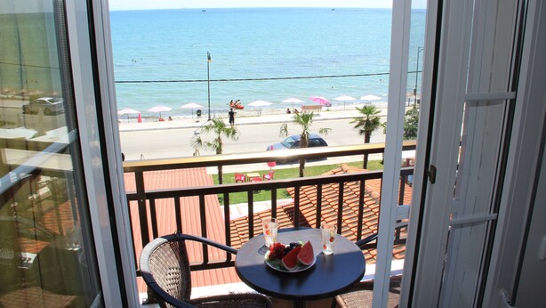 Un balcon avec une table dressée, ornée de fruits et de boissons, donnant sur une plage parsemée de parasols et de palmiers.