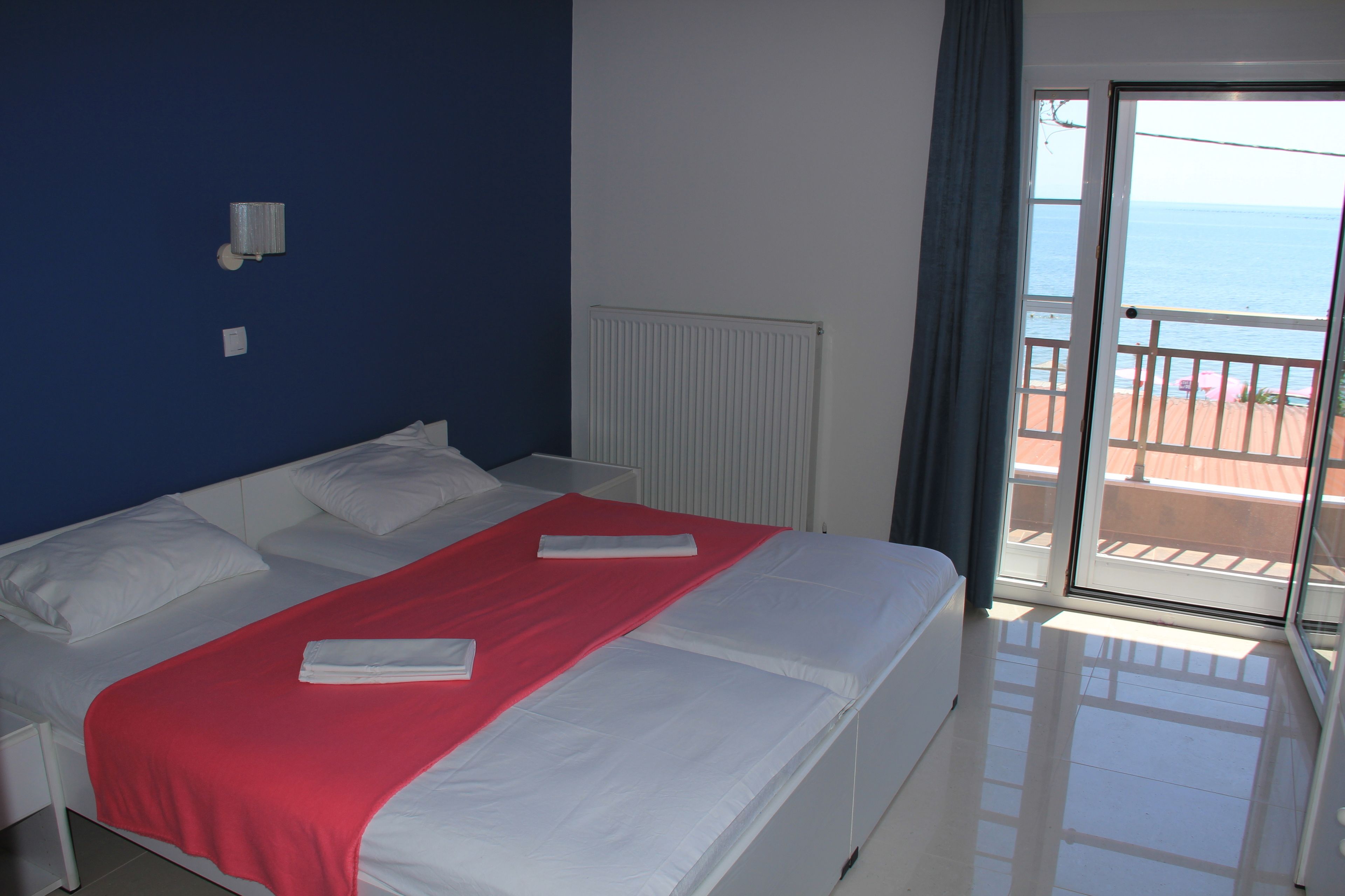 A hotel room with a bed, a red blanket, white pillows, a white radiator, and a view of the sea through a sliding glass door.