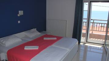 A hotel room with a bed, a red blanket, white pillows, a white radiator, and a view of the sea through a sliding glass door.