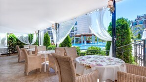 An outdoor patio with wicker furniture, a round table with a floral tablecloth, and a view of a swimming pool and buildings in the background. - Apart-hotel Dawn Park (Sunny Beach)