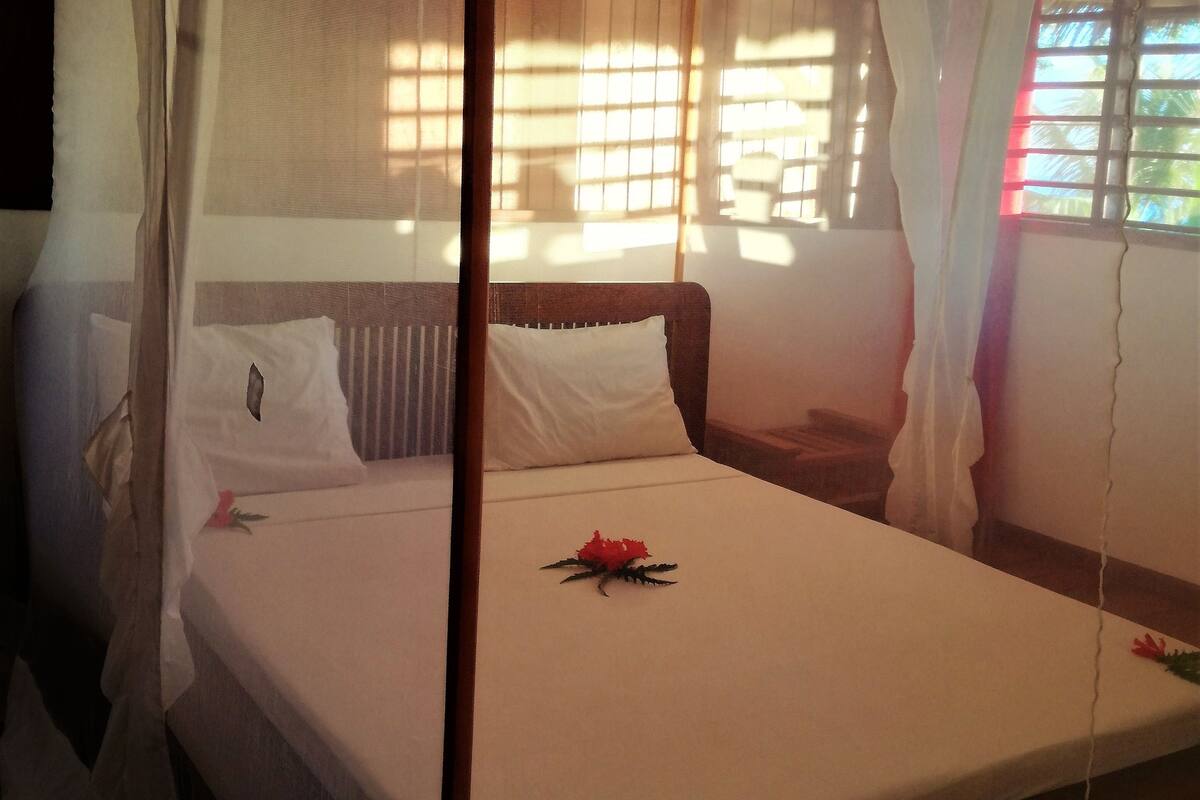 Un lit à baldaquin, des draps blancs et une seule fleur rouge posée sur le lit.