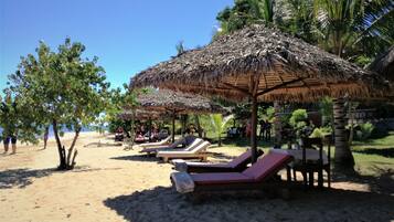 A beach with thatched umbrellas, lounge chairs, and palm trees.