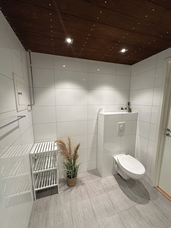 A modern bathroom with white tiles, a wooden ceiling, a toilet, a sink, and a shower area.