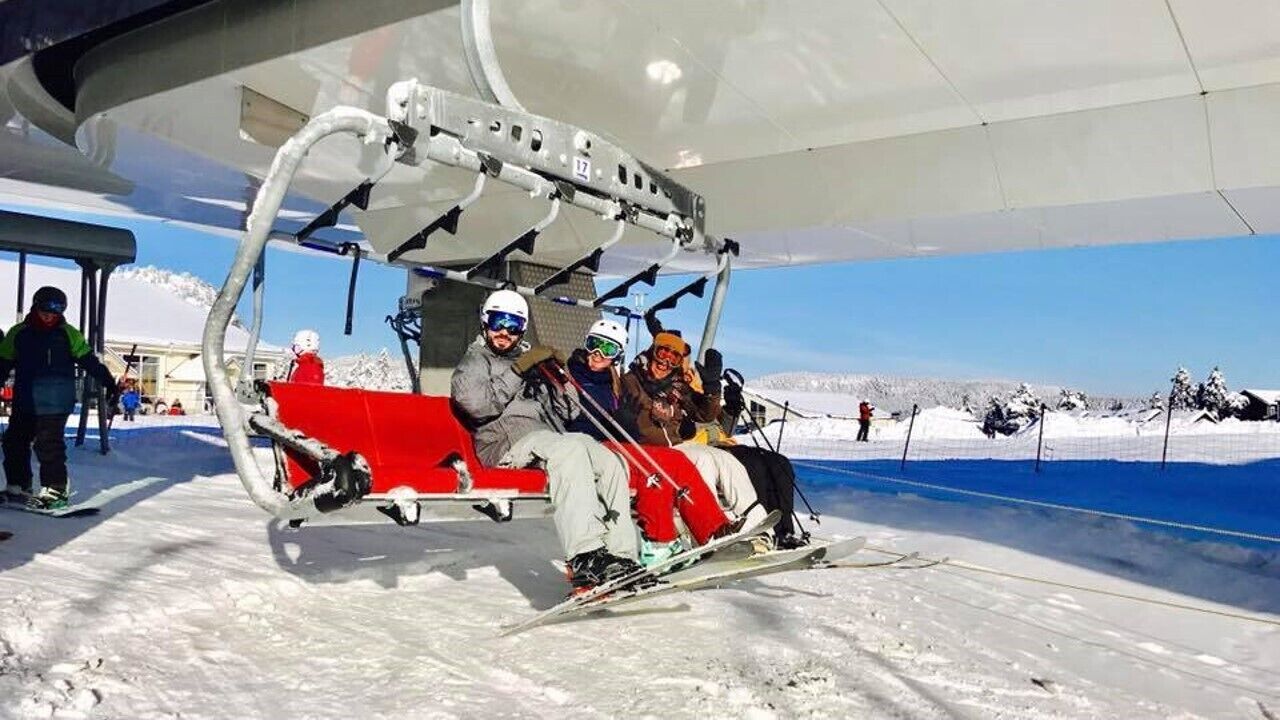 Gondola com passageiros sentados em cadeira vermelha, chão coberto de neve e céu claro ao fundo.