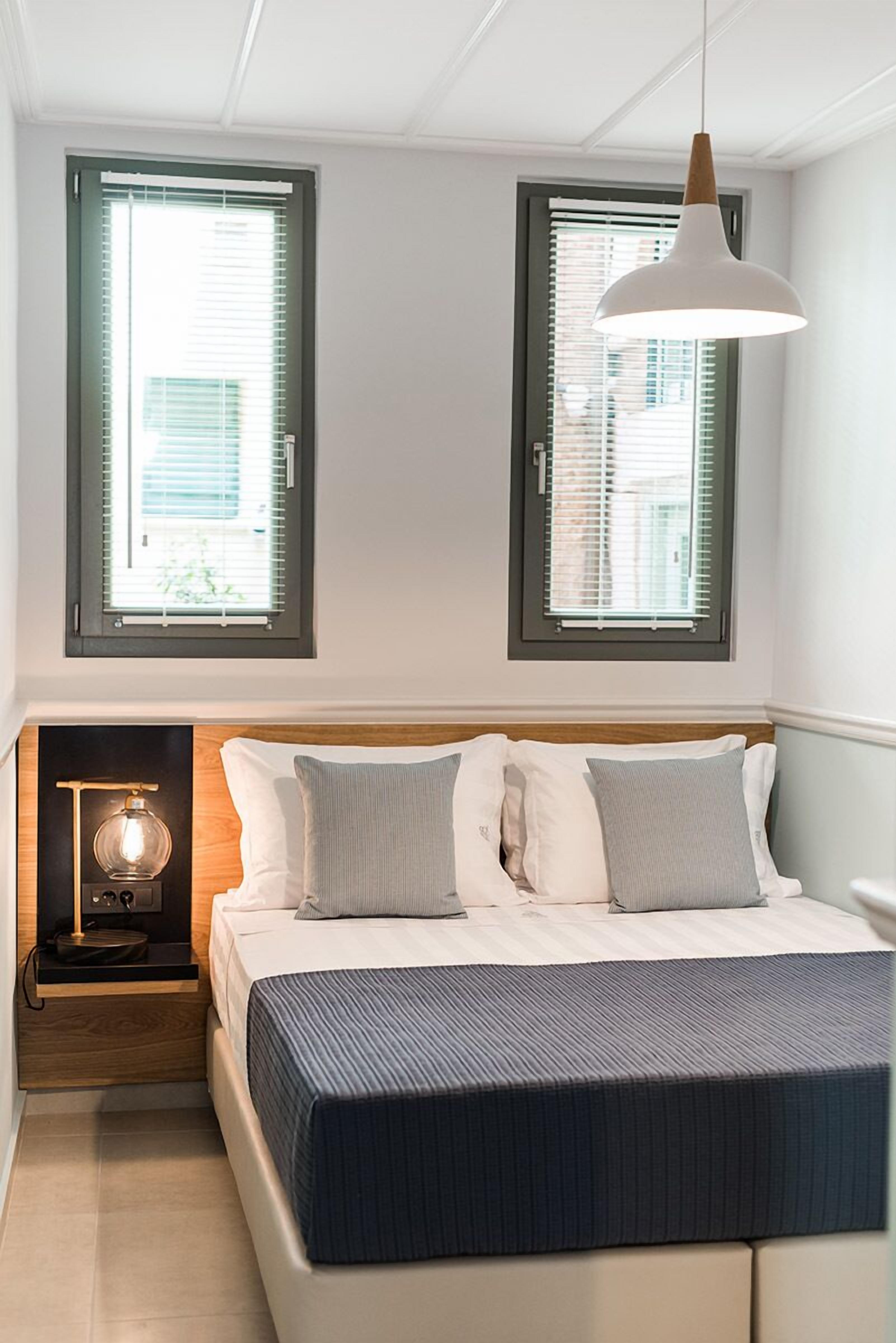 A bedroom with a bed, bedside lamp, and two windows with blinds.