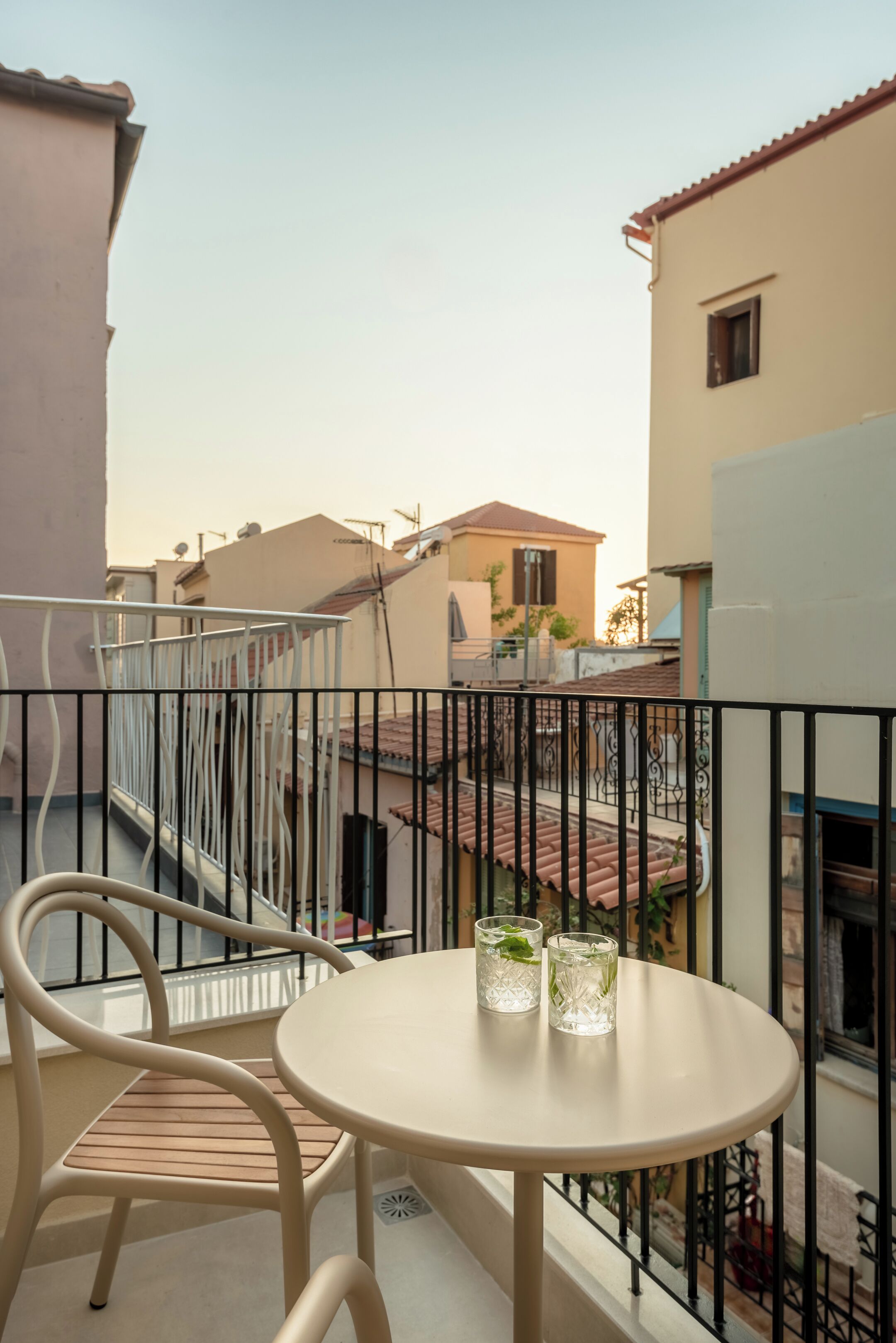 A balcony with a round table and two chairs, overlooking a residential area with houses and a clear sky.