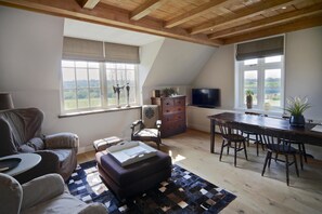A spacious loft with a high ceiling, wooden beams, a dining area with a large table and chairs, a cozy seating area with a sofa and ottoman, and large windows offering a view of the countryside. - Buitenplaats Welsdael (Margraten)