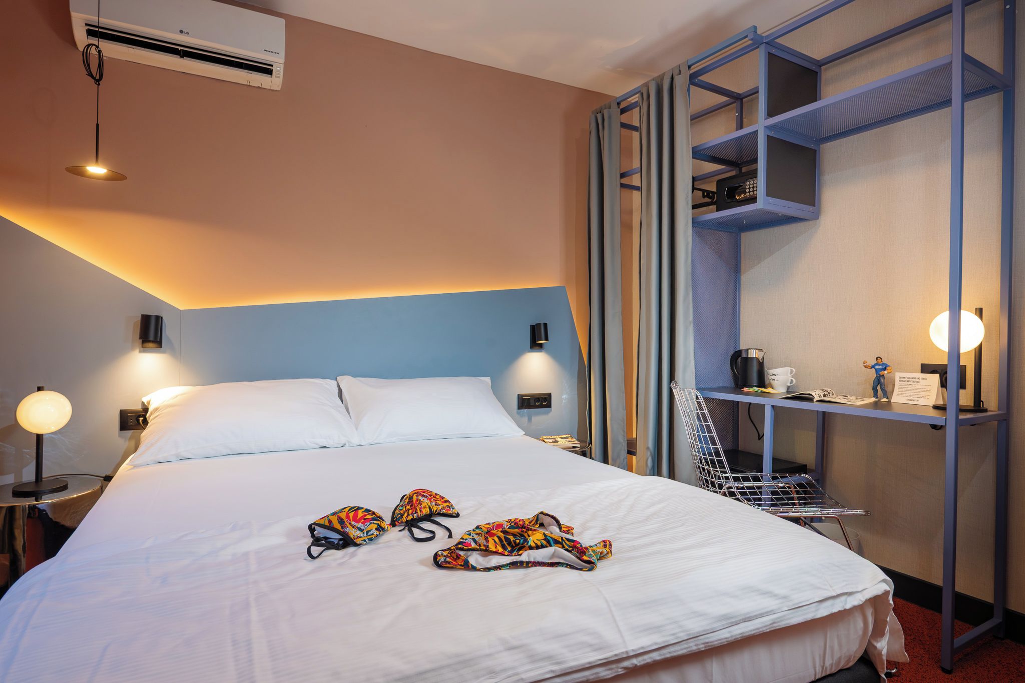 Habitación de hotel con cama, dos lámparas de noche, aire acondicionado mural, estante con tetera y tazas, y una mesita con una figurita.