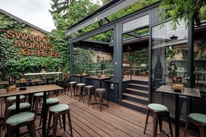 An outdoor seating area with wooden tables and stools, surrounded by greenery and a wall with illuminated letters. - Stay Swanky (Zagreb)