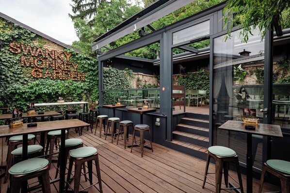 An outdoor seating area with wooden tables and stools, surrounded by greenery and a wall with illuminated letters. - Stay Swanky (Zagreb)